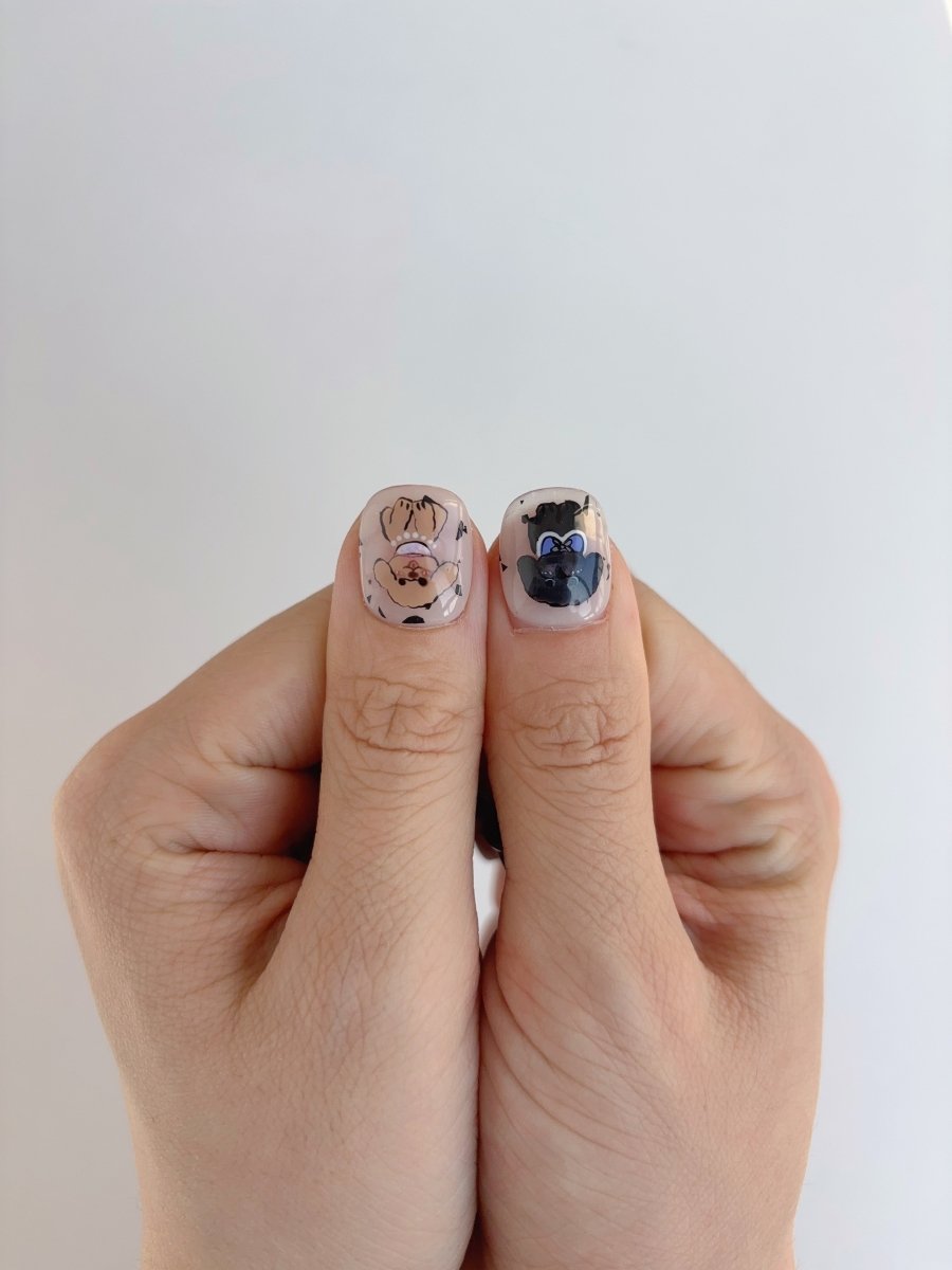 Thumbs showing glossy press-on nails with pug and black dog faces in soft pastel and black designs