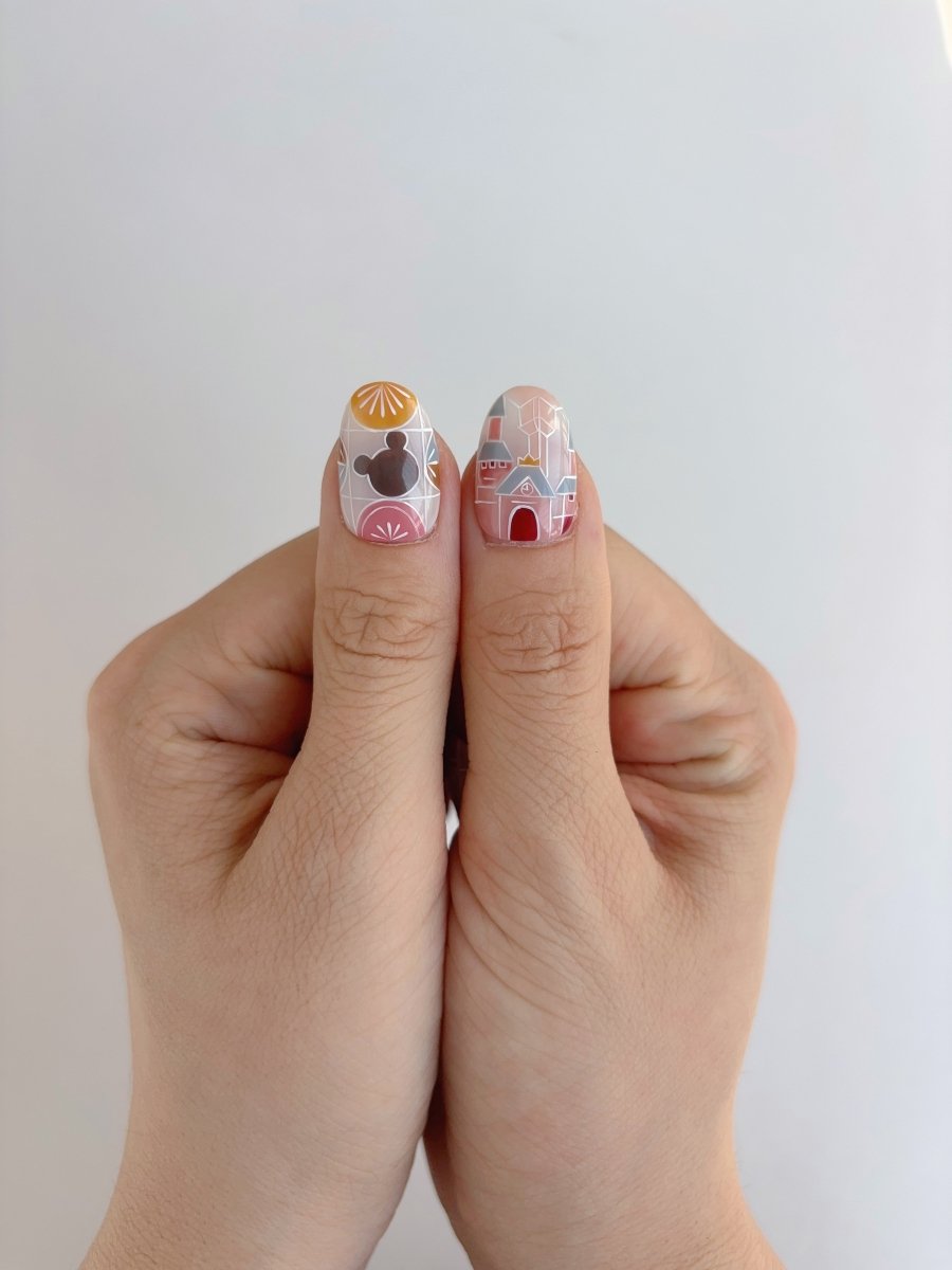 Thumbs displaying press-on nails with pastel pink castle art and orange sunburst mouse silhouette