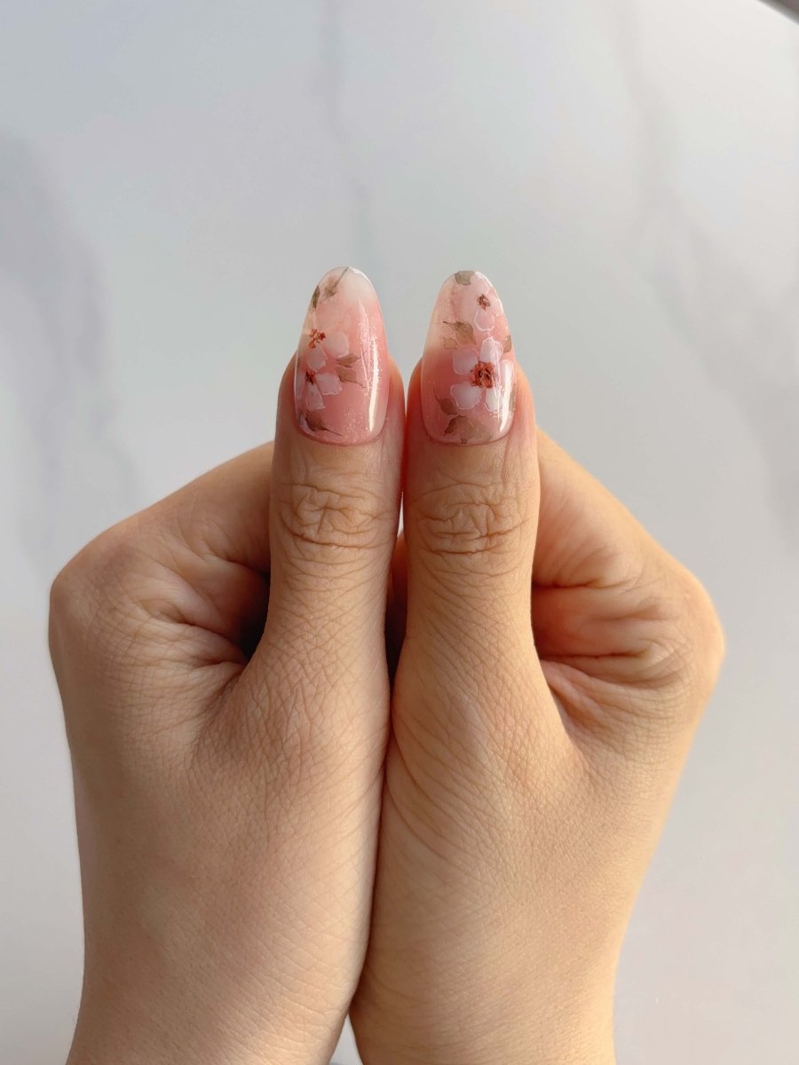 Two thumbs with Sakura Whisper nails featuring delicate cherry blossoms and glossy nude gradient
