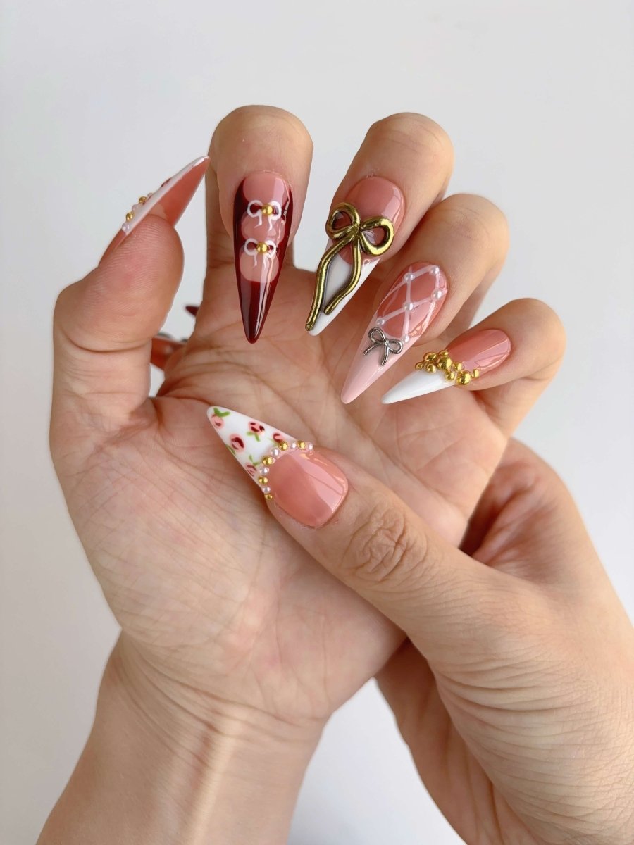 Hand holding long stiletto press-on nails with gold 3D bow, wine red tips, white lattice accents, and pink floral French designs