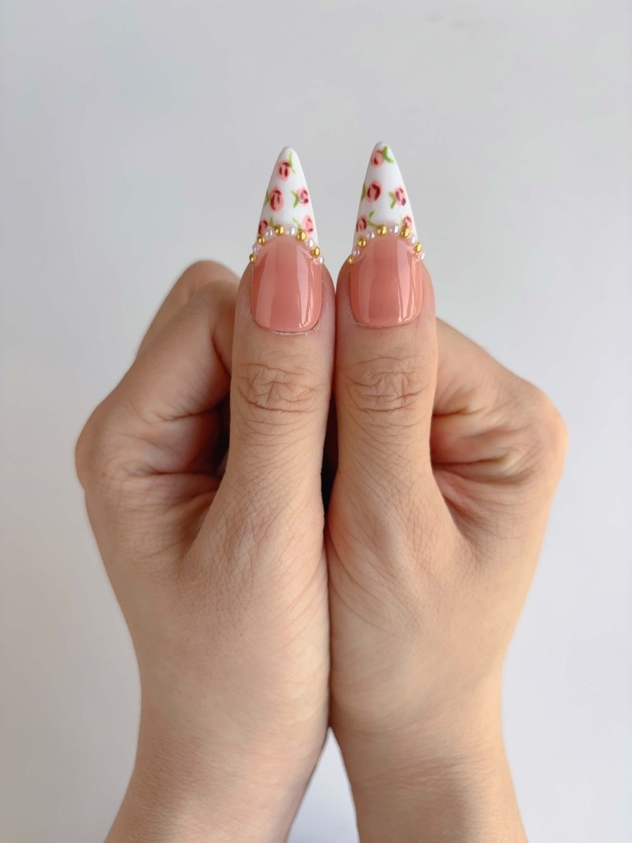 Close-up of thumbs showing white French tips with tiny hand-painted pink roses and gold bead accents