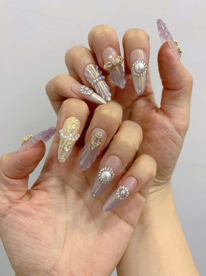 Hand wearing Royal Shell nails with gold charms, pearls, and crystal accents